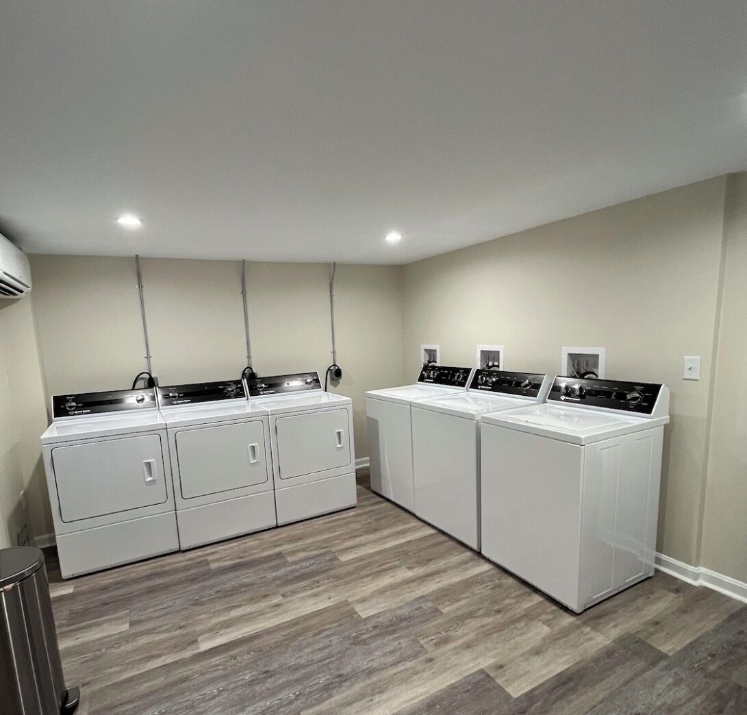 A laundry room with four stacked washer and dryer units, a trash can, and a ceiling-mounted air conditioning unit.