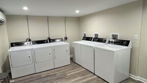 A laundry room with four stacked washer and dryer units, a trash can, and a ceiling-mounted air conditioning unit. - Oceanview! Cozy Condo with Kitchen, Living Area, Porch, 2 Reserved Parking Spots (Old Orchard Beach)