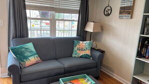 A living room with a grey sofa, two decorative pillows, a teal side table with books and a remote control, and a bookshelf with books and a framed quote. - Oceanview! Cozy Condo with Kitchen, Living Area, Porch, 2 Reserved Parking Spots (Old Orchard Beach)