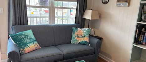 A living room with a grey sofa, two decorative pillows, a teal side table with a book and remote, and a bookshelf with books and a framed quote.