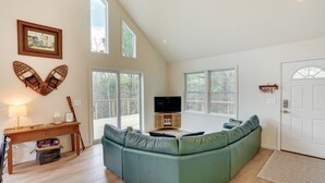 Amplia sala de estar con un sofá seccional verde, una mesa de centro de madera, un televisor sobre un mueble bajo y ventanas grandes que ofrecen vistas al exterior.