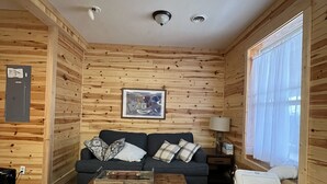 Une pièce confortable, aménagée comme un chalet, avec des murs et des planchers en bois, un canapé sombre agrémenté de coussins à motifs, une table basse en bois et une petite lampe de table blanche.