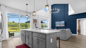 Een moderne keuken met een groot eiland, een zithoek met een grijze bank en uitzicht op een groen landschap door grote ramen.