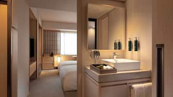 Quarto de hotel moderno com uma cama grande, mesa de cabeceira, televisão e banheiro com pia, espelho e amenities.
