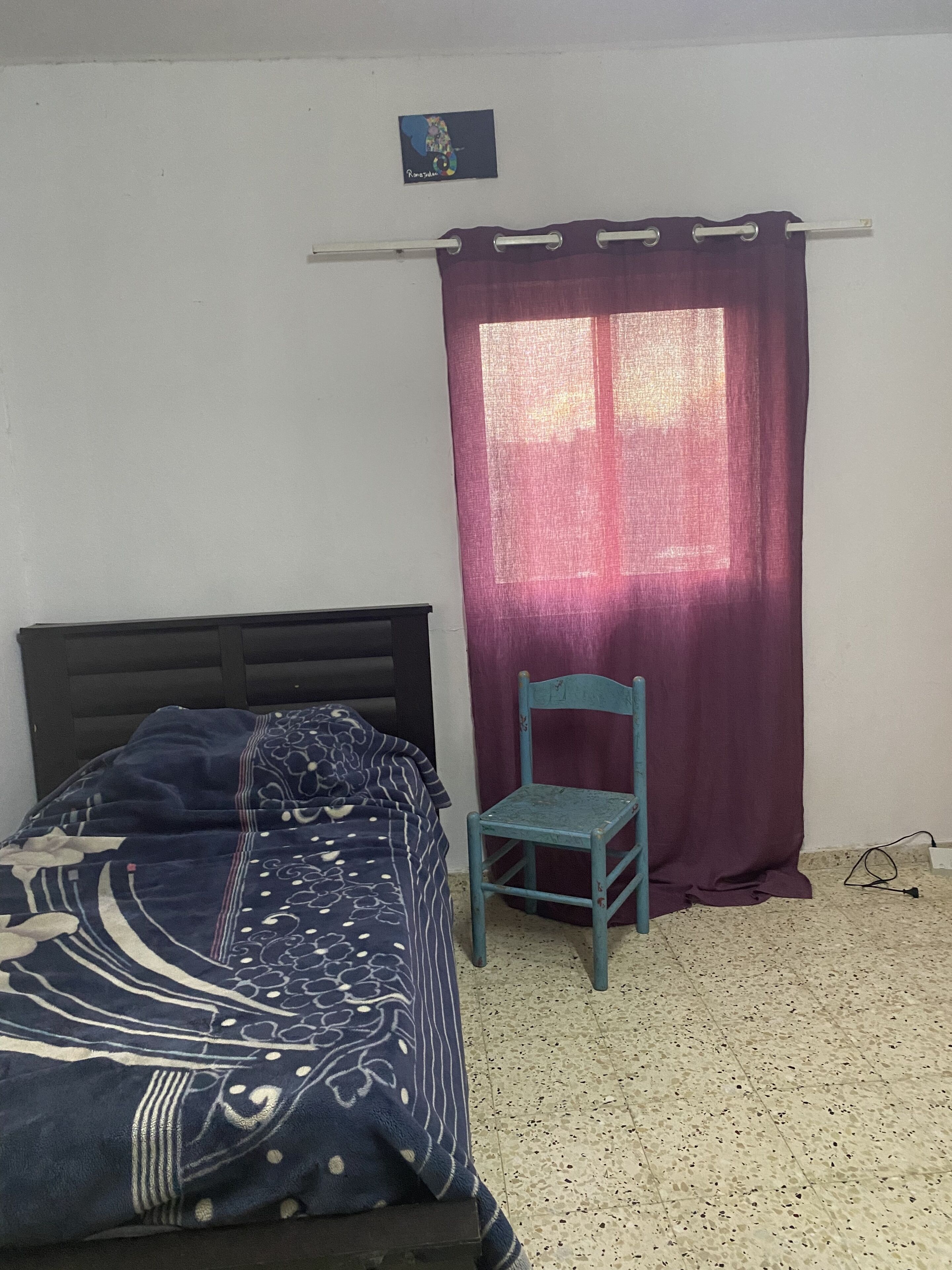 A bedroom with a bed, a chair, a window with curtains, and a small picture on the wall.