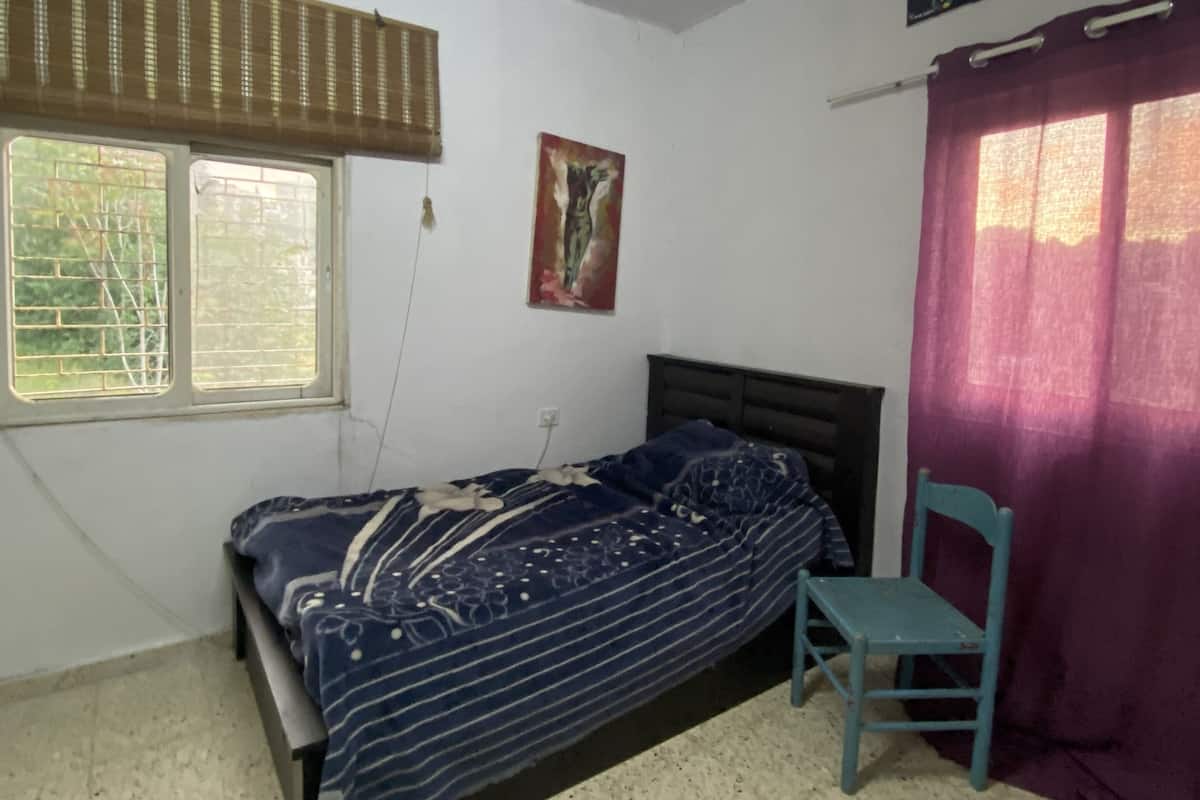 A small, single-bed room with a window, a chair, and a bed with a patterned bedspread.