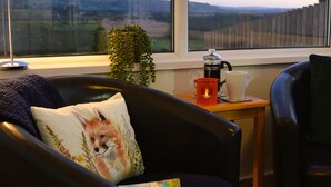 Tunnelmallinen huone, josta avautuu näkymä maaseudulle. Huoneessa on kahvinkeitin, punainen kynttilä ja nojatuolilla kirja.