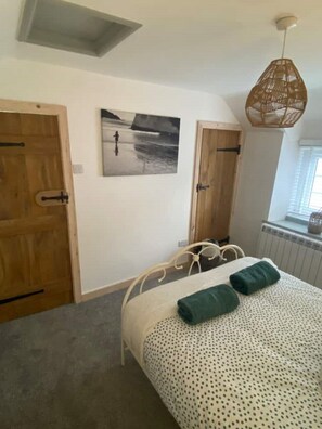 Une chambre à coucher avec un lit, deux portes en bois, un tableau accroché au mur, un luminaire suspendu et une fenêtre donnant sur un paysage.