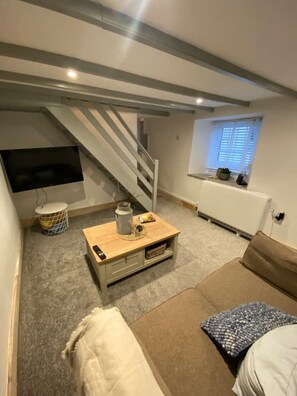 Un espace de vie compact comprenant un escalier, un téléviseur fixé au mur, une petite table avec une lampe et un canapé avec des coussins.