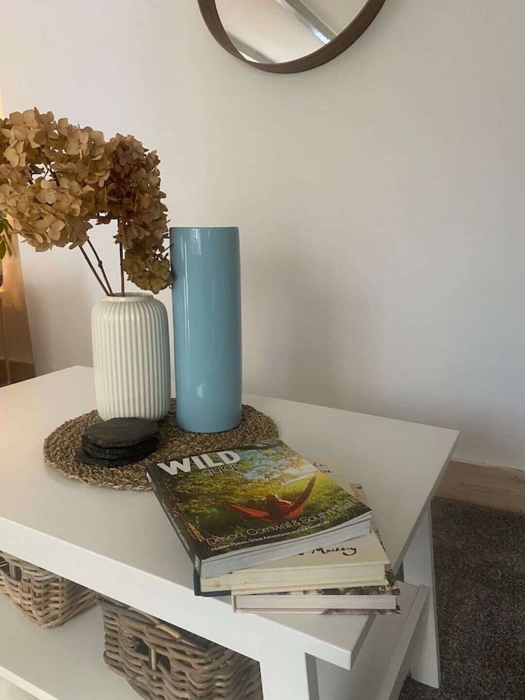 A white table with a blue vase, a book titled 'Wild Guide', and a vase with dried flowers.