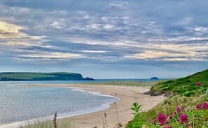A coastal landscape with a sandy beach, grassy hills, and a body of water under a cloudy sky. - Cute, Cosy, Cornish Cottage (Wadebridge)