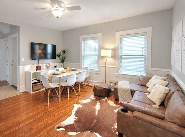 A modern living room with a wooden floor, a brown leather sofa, a dining table with chairs, a TV mounted on the wall, and a ceiling fan. - Luxe apartment at MIT/Harvard with FREE parking (Cambridge)