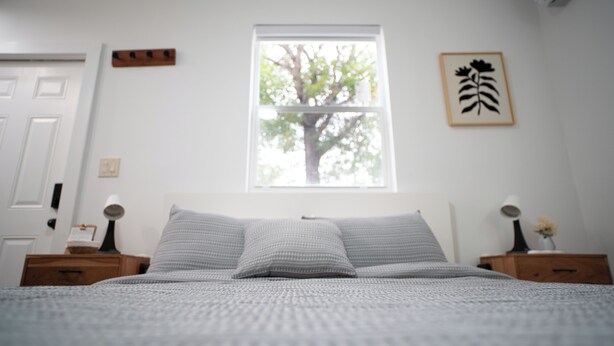 Quarto com cama, duas mesas de cabeceira, janela com vista para uma árvore, um quadro na parede e uma porta.