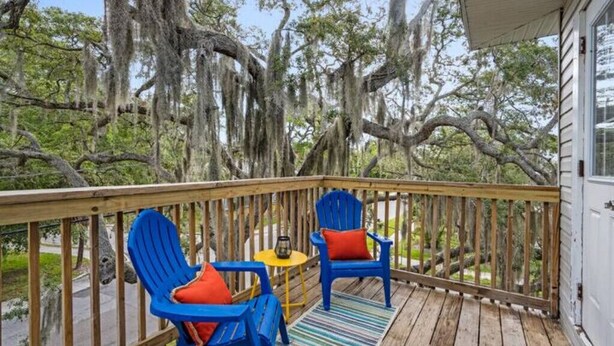 A wooden deck with two blue Adirondack chairs and a small table, overlooking a natural landscape with trees and a building in the background. - Colorful, waterside home (Bradenton)