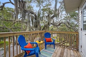 Una terraza de madera con dos sillas Adirondack azules y una mesita, con vista a un paisaje natural con árboles y un edificio al fondo.