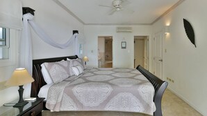 Een slaapkamer met een groot bed, een nachtkastje met een lamp, een plafondventilator en een deur naar een andere kamer.