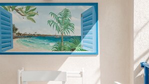 A room with a glass table, white chairs, a painting of a beach scene, and a white wall.