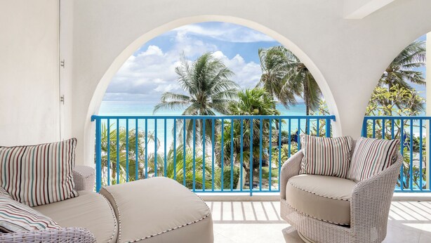 A balcony with a view of the ocean and palm trees, furnished with two wicker chairs and striped cushions.
