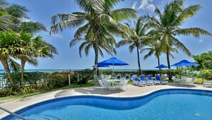 A swimming pool with blue tiles, surrounded by lounge chairs and umbrellas, with palm trees and a view of the ocean.