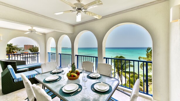 A dining area with a table set for four, overlooking a beach with clear turquoise water and palm trees.