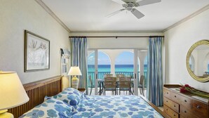 A hotel room with a bed, a dresser, a chair, a table, and a balcony with a view of the ocean.