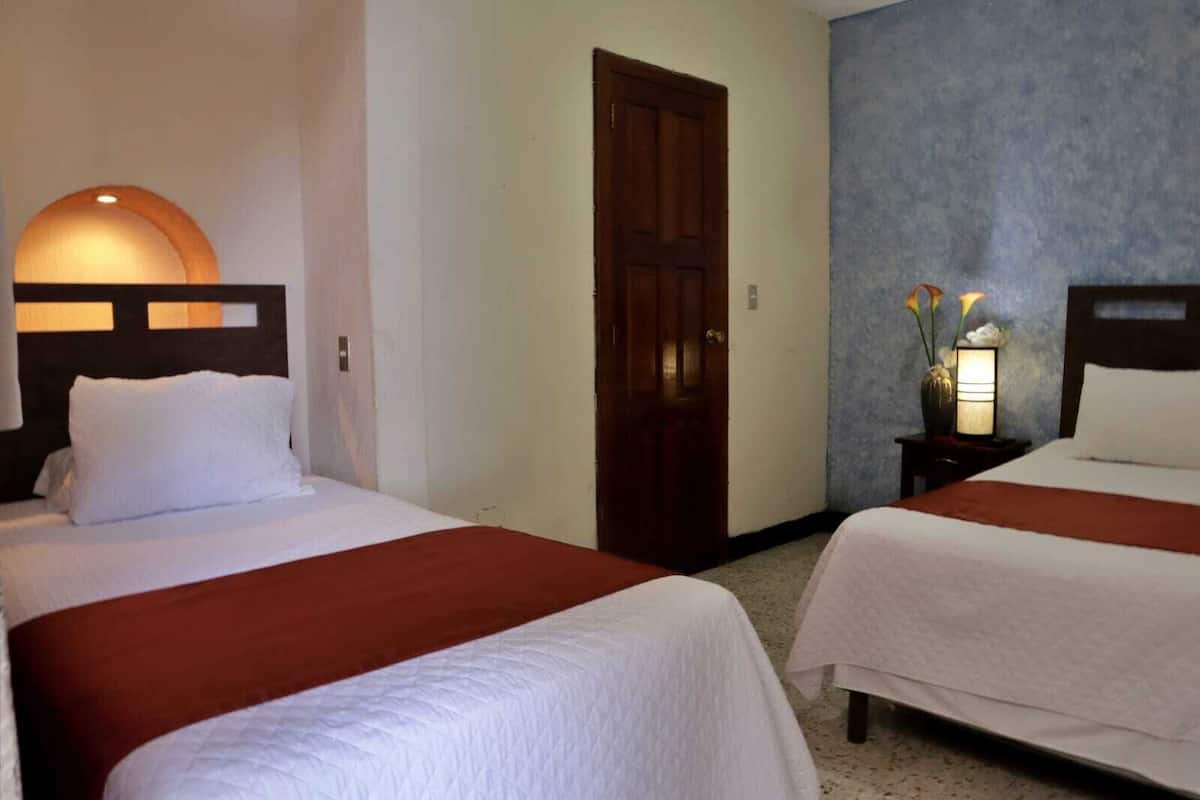 Habitació d'hotel amb dos llits, una porta de fusta, una tauleta de nit amb una làmpada i una paret amb un relleu decoratiu.