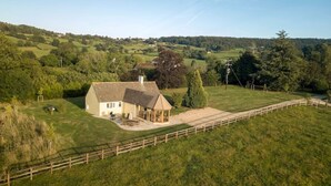 Une maison de campagne avec une cour clôturée, un petit jardin et une vue sur des collines verdoyantes à l’arrière-plan.