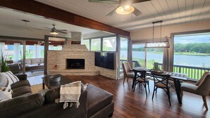 Amplia sala de estar con chimenea de piedra, televisor de pantalla plana, ventilador de techo y gran ventana con vista a un lago.