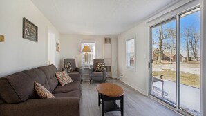 A living room with a brown sofa, a small round table, and a gray armchair with patterned cushions. There is a window with a view of a snowy outdoor area. - Beautiful 2 bed cottage by Coldwater Lake! (Coldwater)