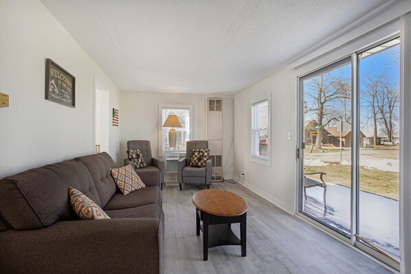 A living room with a brown sofa, a small round table, and a gray armchair with patterned cushions. There is a window with a view of a snowy outdoor area.