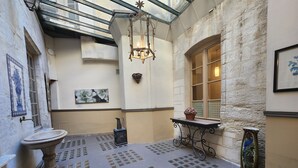 Un pasillo estrecho con pared de piedra, suelo de baldosas, una mesita con un objeto decorativo, una lámpara fijada a la pared y una ventana con vistas.