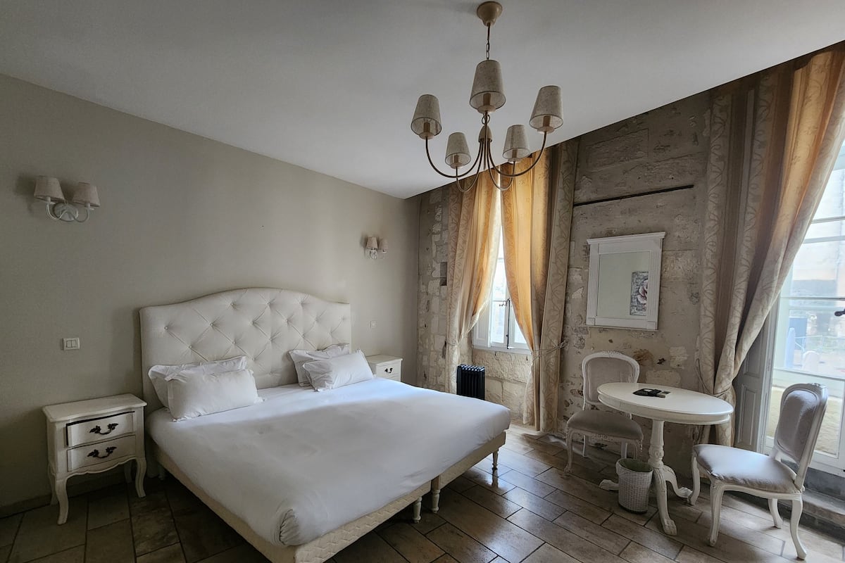 Un dormitorio con una cama grande, mesitas de noche, una araña, una pequeña mesa redonda con sillas y una ventana con cortinas.