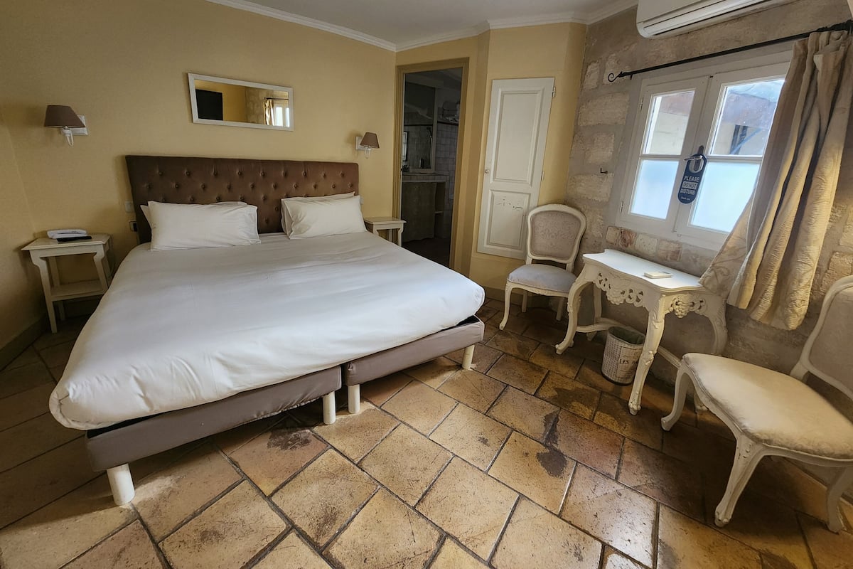 Habitación de hotel con una cama grande, una silla, una mesita, un espejo y un equipo de aire acondicionado.