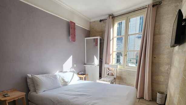 A bedroom with a bed, a bedside table, a mirror, a window with curtains, and a wall-mounted TV. - Hôtel du Palais des Papes (Avignon)