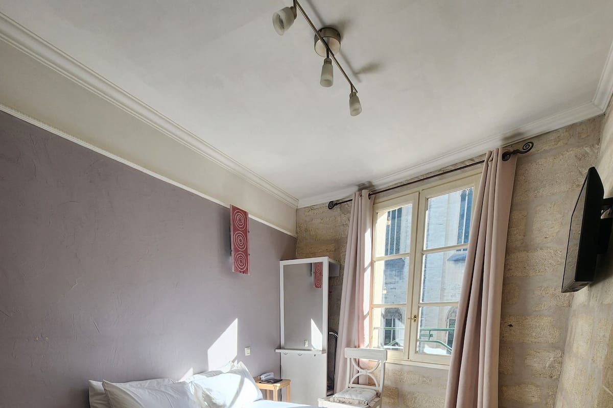 Un dormitorio con cama, mesita de noche, espejo, ventana con cortinas y televisor montado en la pared.