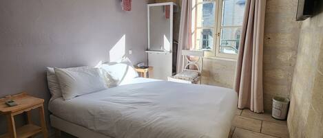 A bedroom with a bed, a bedside table, a mirror, a window with curtains, and a wall-mounted TV.