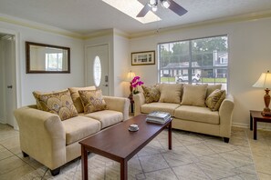 A living room with two beige sofas, a wooden coffee table, a large window with blinds, a ceiling fan, and a framed picture on the wall. - Savannah Vacation Rental ~ 8 Mi to River Street! (Savannah)