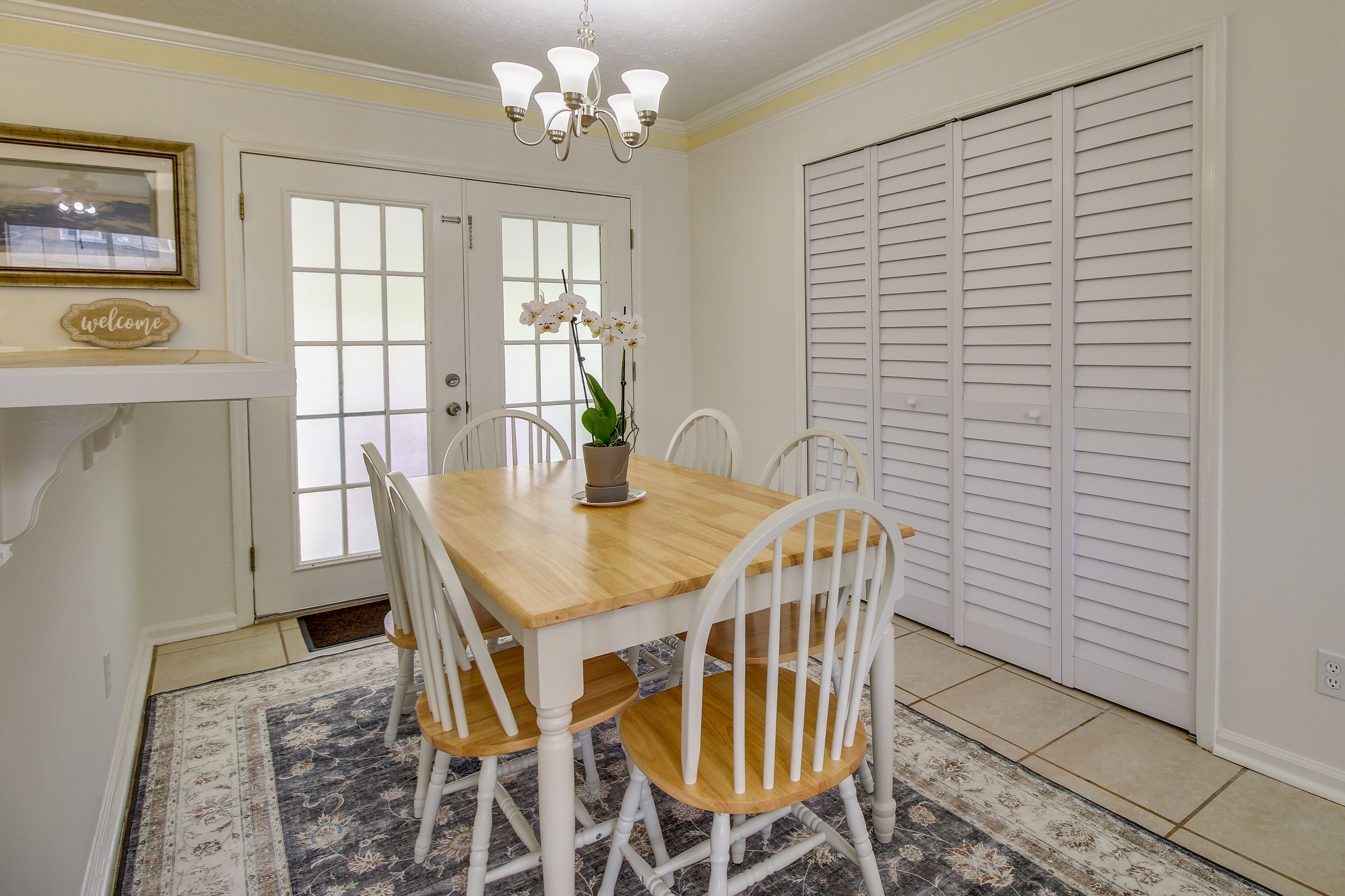 Un comedor con mesa de madera y sillas blancas, una araña y una puerta con paneles de vidrio esmerilado.