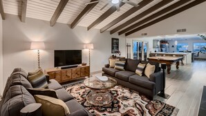 A spacious living room with a large sofa, a coffee table, and a television. There is a ceiling fan, a fireplace, and a dining area in the background.