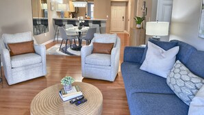 A modern living room with two grey armchairs, a blue sofa, and a coffee table with books and remotes. A dining area with a chandelier and a kitchen in the background is visible. - 3 Pools, Hot Tub, Sports Courts, Resort, Golf, Gorgeous All New Condo! (Branson)