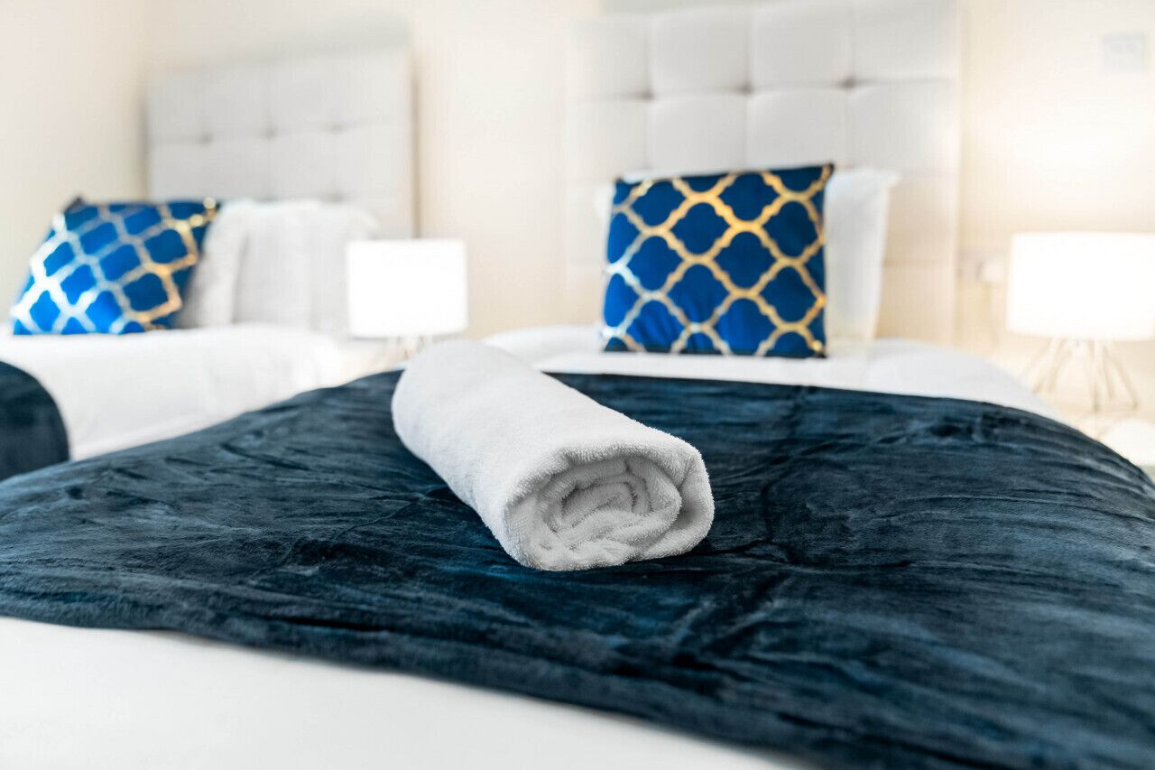 Una cama bien tendida con una toalla blanca enrollada, una colcha azul oscuro con textura y dos almohadas con diseños azules y dorados.