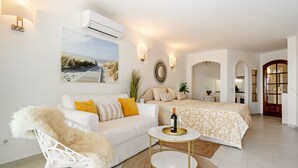 A modern bedroom with a white sofa, a bed with a patterned duvet, a small round table with a bottle and glasses, and a wall-mounted air conditioner. - WELCS ESTUDIO 057 EMP Frente al Mar (Empuriabrava)