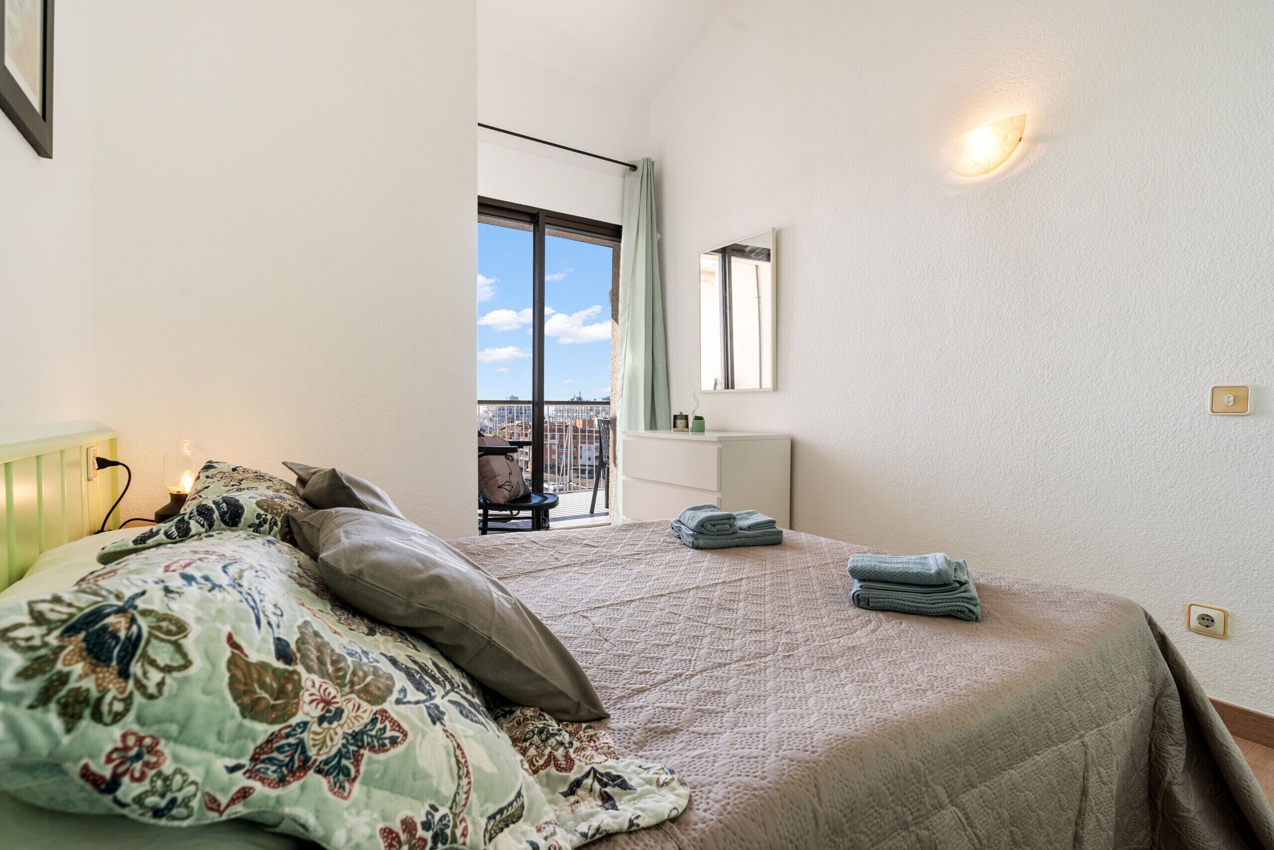 A bedroom with a large bed, a window with a view, a bedside table, and a wall-mounted light.
