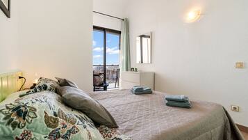 A bedroom with a large bed, a window with a view, a bedside table, and a wall-mounted light.