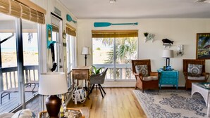 Ein geräumiges Wohnzimmer mit großem Fenster zum Strandblick, Holzboden und verschiedenen Sitzgelegenheiten, darunter ein Sessel, eine Couch und ein kleiner Tisch.