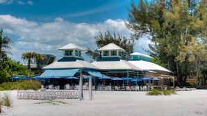 A beachside restaurant with outdoor seating and covered areas, surrounded by trees and a clear sky.