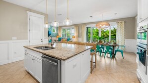 A kitchen with a central island, white cabinetry, and a stainless steel dishwasher. There is a dining area with teal chairs and a table, and a large window with curtains.