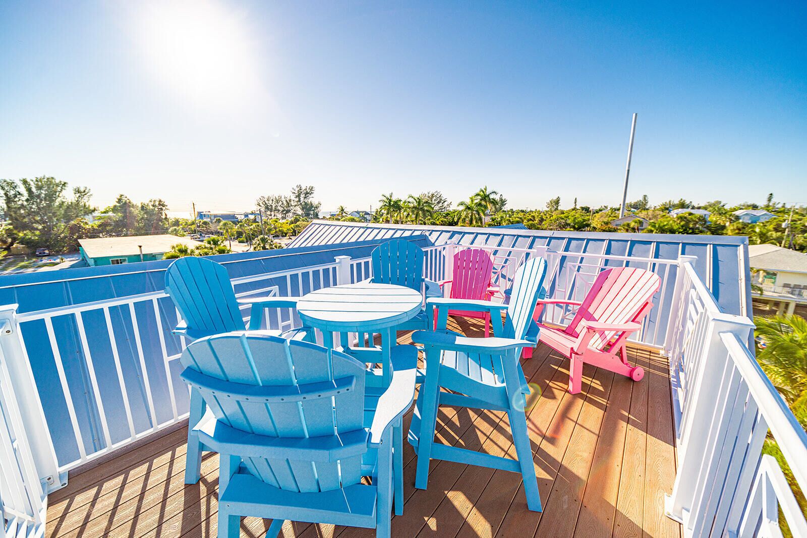 Una terraza con sillas Adirondack azules y rosadas, una mesa redonda y una barandilla blanca.