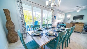 A dining area with a table set for six, surrounded by teal chairs, a chandelier, and a large window with a view of palm trees.