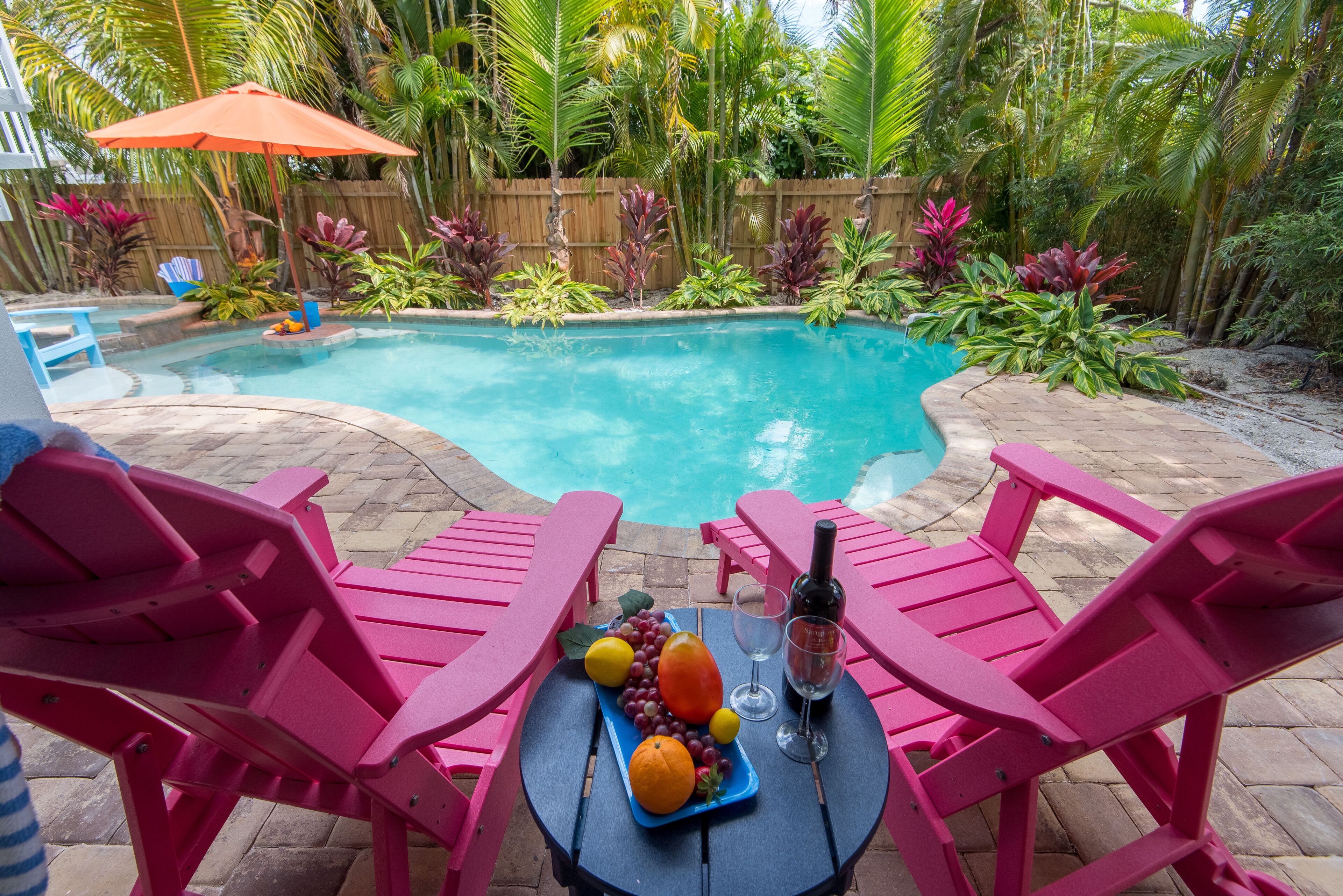 En baghave med en swimmingpool, omgivet af tropiske planter og et træhegn. Der er pink farvede stole og et bord med frugt og en flaske.
